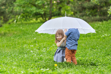 little girl and boy are hiding under an umbrella. brother and sister are playing in the park.