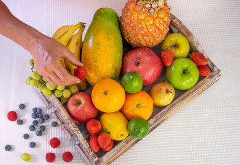 Wooden basket full of fresh fruit on a white background. Citrus fruits, papaya, pineapple, banana, pear, apple, strawberry, pomegranate, blueberry. Female hand holding a strawberry