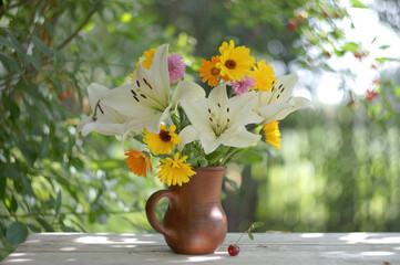 Still life with a cheerful bouquet of white lilies and bright yellow calendula flowers in a brown clay jug on a white table top in a sunny summer garden