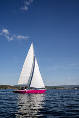 Fototapeta premium A crimson yacht with raised white sails sails in the middle of the lake. People are resting on the deck.