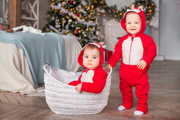 twins toddlers in red santa's reindeer suits sit next to each other at home against the background...