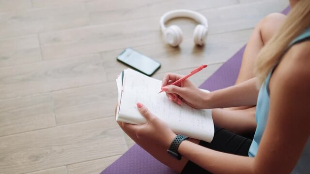 Side view of woman writing something in notepad from phone while sitting on the yoga mat