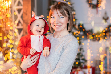 happy mom playing with her toddler in red santa reindeer costume on christmas tree background
