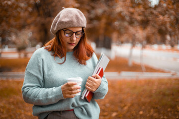 Young woman student in glasses with hot coffee drink walks in the autumn park