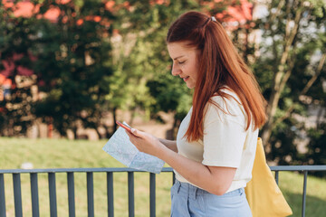 Naklejka premium Attractive young female tourist is exploring new city. Happy redhead Woman with map and smartphone in hands lost in new place, relaxation or traveling concept
