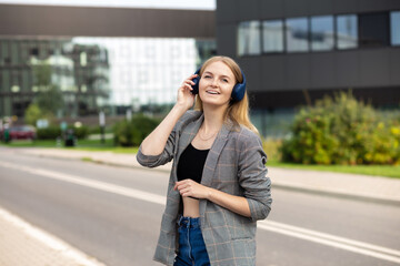 Portrait of young happy woman listening to music with headphones and smiling while walking on the street in the city. Music lover enjoying music. Portrait of businesswoman walking and smiling outdoor