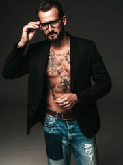 Obraz premium Portrait of handsome confident stylish hipster lambersexual model. Sexy modern man dressed in black jacket. Naked torso with tattoos.Fashion male posing in studio on dark background. In spectacles