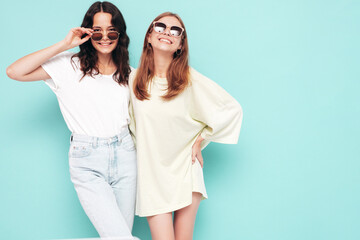 Two young beautiful smiling brunette hipster female in trendy summer clothes. Sexy carefree women posing near blue wall. Positive models having fun. Cheerful and happy. In sunglasses