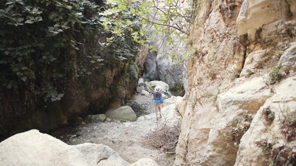 Back view of young woman in blue blouse, denim shorts and white hat walking along canyon of high rocks during warm summer day. Adventure of young female person. Travelling concept.