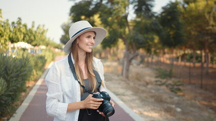 Young woman photographer wanderer. Happy young woman using modern digital camera for taking photos while walking. Female tourist in summer hat and clothes saving memories from trip