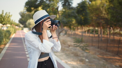 Young woman photographer wanderer. Happy young woman using modern digital camera for taking photos while walking. Female tourist in summer hat and clothes saving memories from trip