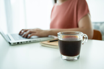 Beautiful young Asian woman holding a cup of hot black coffee on the table while working. Asian young woman enjoy drinking a hot coffee in the morning before starting online meeting from home.