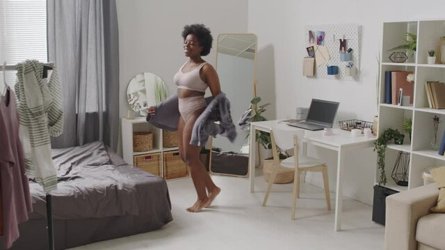 Full-length slowmo shot of young confident plus size African-American woman in casual pink underwear dancing at bedroom alone spending time with herself