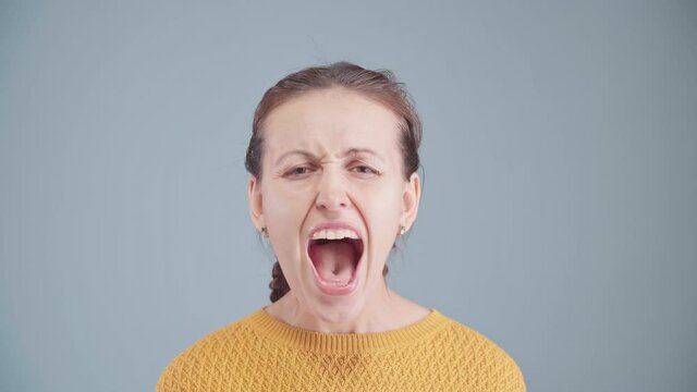 A beautiful middle-aged woman screams with her mouth wide open and looking at the camera. Blue background