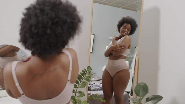 Rear-view medium slowmo shot of smiling young curvy African-American woman in underwear enjoying her reflection standing in front of mirror at bedroom. Concept of self-love and body positivity