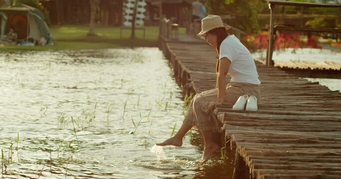 A female tourist in a brown hat, white T-shirt and brown skirt sits on a wooden bridge by the lake, looking happily at the waves, use her feet to swing the water relaxing on vacation at sunset time.