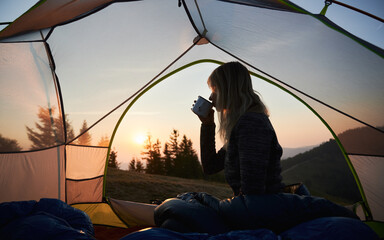 Silhouette of blonde woman drinking hot tea from metal cup, relaxing in her tent and admiring dawn...