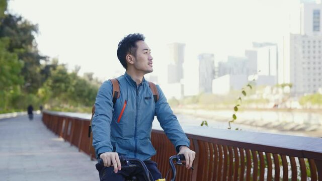 young asian man riding bicycle on riverfront path looking at view