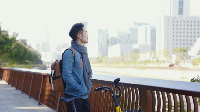 young asian man standing by the river looking at view while commuting by bike