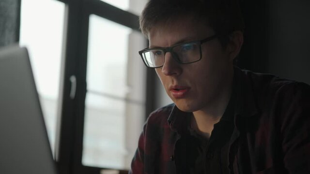 A young specialist works in a bright office with a laptop, mumbles, moves lips and types on a laptop. Coworking. The student is studying, working on a project. Portrait.