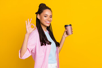 Photo of young happy positive dark skin woman make okay sign hold coffee cup wear striped shirt isolated on yellow color background