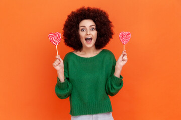 Excited woman with Afro hairstyle in green casual style sweater holding two candies with heart shape, looking at camera with happy positive expression. Indoor studio shot isolated on orange background