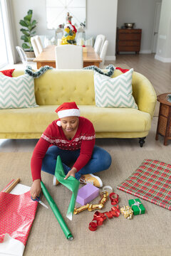 Happy african american plus size woman in santa hat wrapping presents at home