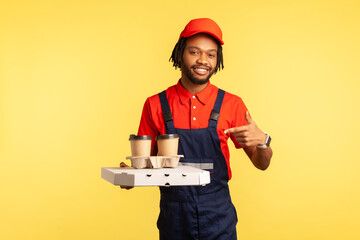 Delivery service. Optimistic courier man in blue overalls pointing at coffee and box with fast food, offering drinks in disposable cups and pizza. Indoor studio shot isolated on yellow background.