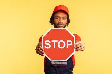 Portrait of serious handyman wearing uniform and red visor cap looking at camera, holding out showing red stop traffic sign, warns of danger. Indoor studio shot isolated on yellow background.