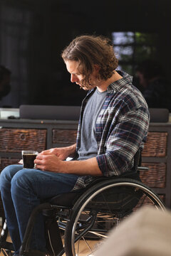 Caucasian disabled man sitting on wheelchair holding coffee cup reading a book at home