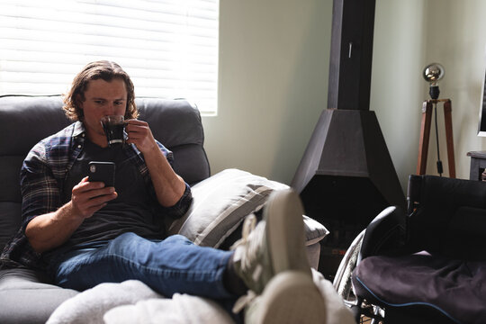 Caucasian disabled man drinking coffee and using smartphone sitting on the couch at home