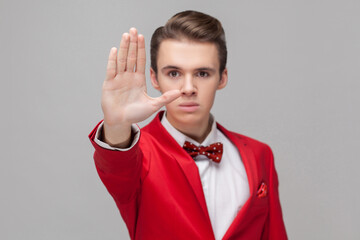 Stop! Portrait of confident gentleman with stylish hairdo in red tuxedo and bow tie showing prohibition hand gesture, saying no, forbidden, deny concept. indoor studio shot isolated, gray background