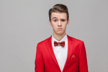 Portrait of confused young man with stylish hairdo in red tuxedo and bow tie looking with surprised and puzzled face, thinking what is going on. indoor studio shot isolated on gray background