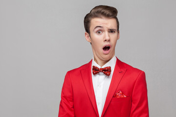 Portrait of funny surprised man with stylish hairdo in red tuxedo and bow tie staring in surprise at camera with comical dumb grimace, making a face. indoor studio shot isolated on gray background