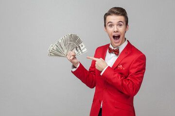 Wow, look at my money! Portrait of surprised man with stylish hairdo in red jacket and bow tie pointing at bunch of dollars, expressing extreme joy and amazement. indoor studio shot, gray background