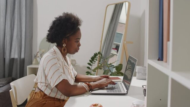 Side-view medium slowmo shot of young attractive African-American woman in casualwear working on laptop sitting by table at cozy apartment