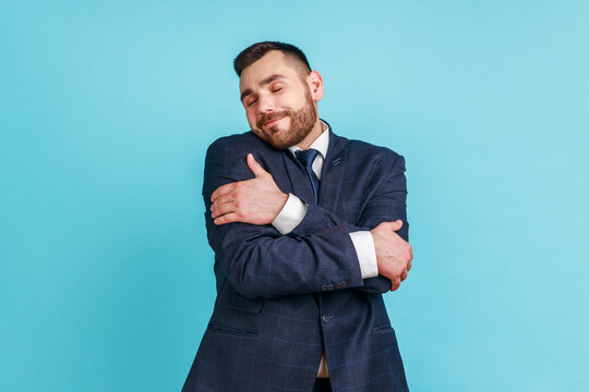 I love myself! Portrait of narcissistic selfish young adult man in suit embracing herself, closed eyes and smiling with pleasure, complacency and egoism. Indoor studio shot isolated on blue background