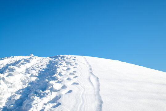The top of a snow-covered hill with trampled path and footprints against the blue winter sky