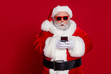 Fototapeta na wymiar Portrait of attractive bearded amazed Santa holding in hands using device gadget isolated over bright red color background