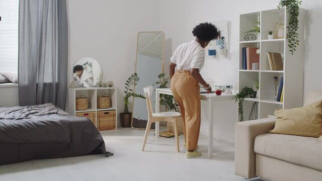 Full-length slowmo shot of young beautiful plus size African-American businesswoman in casualwear sitting down at table in bright modern apartment and starting work on laptop