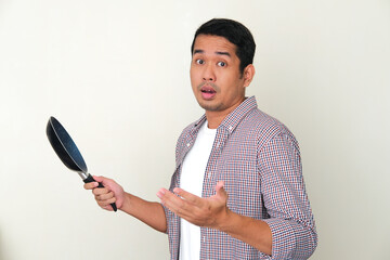 Adult Asian man showing confused face expression while holding frying pan