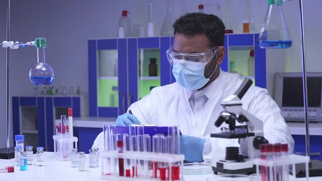 Indian scientist writing on clipboard near test tubes and vaccines in lab