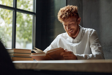 Young redhead man reading book in modern library