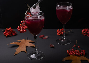 A piece of ice in the form of a skull falls into a glass with a red drink, a hanging bunch of rowan berries, around autumn leaves on a gloomy background. Halloween drinks concept.