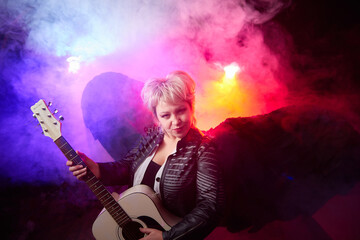 Fototapeta na wymiar Self-assured adult blonde middle age woman with black angel wings and guitar like a rock musician. Model posing in studio on black background. Dangerous cupid on Valentine's day