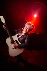 Obraz premium Self-assured adult blonde middle age woman with black angel wings and guitar like a rock musician. Model posing in studio on black background. Dangerous cupid on Valentine's day