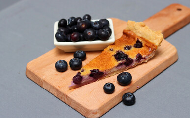 Slice of Blueberry tart on wooden cutting board. Sweet blueberry cake on a table. Homemade dessert close up photo. Healthy eating concept. Grey background with copy space. 