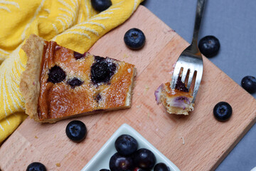 Mustikkapiirakka - Finnish Blueberry pie. Top view photo of tart in a baking form, slice of blueberry cake, fork, kitchen towel and cutting board on a table. 