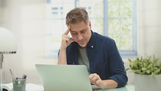 Happy man with laptop computer at home. Businessman working at desk in home office. Portrait of mature age, middle age, mid adult man, bearded, smiling, thinking, authentic look.