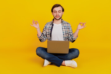 Full size photo of a happy young man holding laptop meditate yoga om while sitting on a floor isolated over yellow color background